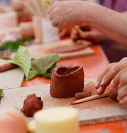 Hände an einem Tisch, die aus rotbraunem Ton Gefäße formen und mit Hilfsmitteln wie kleinen Holzstäben oder Blättern bearbeiten