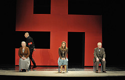 Die Schauspieler/innen Hellena Büttner, Volker Jeck, Katrin Reuter, Peter Bause sitzen in einer Szene nebeneinander auf Stühlen . Im Hintergrund ein riesiges Hakenkreuz in Rot.
