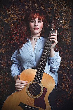 Zu sehen ist die Singer-Songwriterin Jenny Bright. Sie langes, rotes, lockiges Haar und trägt eine hellblaue Bluse. Vor sich in der Hand hält sie eine Akustik-Gitarre. Im Hintergrund ist Laub in Herbstfarben zu sehen, es scheint als liege sie auf einem Bett aus Blättern.