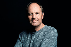 Der Autor Jens Rosteck ist abgebildet und trägt einen bläulichen Strickpullover vor schwarzem Hintergrund. Er lächelt in die Kamera. 