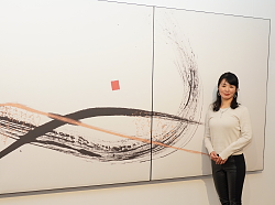 Die Künstlerin steht vor einem großen dreiteiligen Werk, das eine schwungvolle Kalligrafie zeigt. 