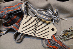 Auf grauem Tuch sind bunte Bänder und ein hölzerner Webkamm dargestellt.