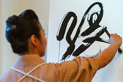 Die Kalligrafriekünstlerin in japanischer Tracht ist von hinten aufgenommen und hat einen Pinsel in der Hand. Sie malt ein japanische Schriftzeichen. 