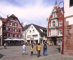 Mehrere Kinder und eine Stadtführerin gehen über einen gepflasterten Platz umgeben von Stadthäusern.