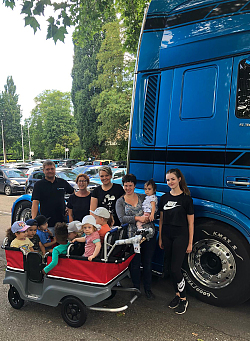 Vor einem blauen Lastwagen, von dem nur die Führerkabine zu sehen ist, stehen von links nach rechts Andreas Schwindt (Schwindt Transporte), Manuela Lackert (stellvertretende Leitung Kita Max-Planck-Straße), Anna Steba, Sabrina Kindle, Gina Siewert (alle Kita Max-Planck-Straße). Davor sitzen neunKinder in einem großen Gefährt, das an einen Wagen einer Achterbahn erinnert. Sie alle tragen Sonnenschutz. 
