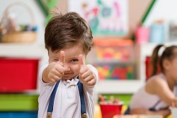 Ein kleiner Junge mit einem weißen T-Shirt, blau-weis-gestreifter Hos und blauen Hosenträger streckt beide Arme nach vorne und zeigt mit den Daumen nach oben. Im Hintergrund sieht man verschwommen Kinder und eine Frau an einem Tisch spielen.