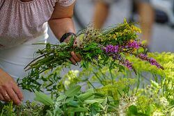 Auf dem Foto sind Frauenhände zu sehen, welche verschiedene Kräuter in grün und violett zusammenschnürt. 