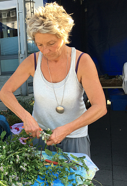 Johanna Harter, gekleidet mit einem ärmellosen Top, steht hinter einem Tisch, auf dem mehrere Kräuter liegen. Sie bearbeitet die Kräuter mit einer Rebschere. 