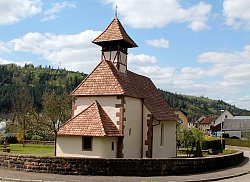 Das Bild zeigt die Galluskirche in Kuhbach. Ein kleines, verputztes Gebäude mit Steinecken, einem viereckigen Erker und Spitzdach auf dem ein kleines Glockentürmchen ist.