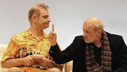 Das Bild zeigt die Schauspieler Heinrich Schafmeister (links) und Leonard Lansink (rechts) auf einem Sofa sitzend. Leonard Lansink hebt belehrend den Zeigefinger, Heinrich Schafmeister schaut mit einem geschlossenen Buch in beiden Händen auf den Finger.