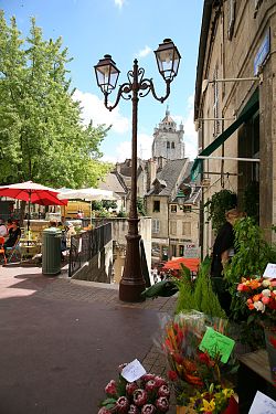 Das Bild zeigt eine mittelalterliche Gasse in Dole. Links sitzen Menschen in einem Café, rechts stehen Blumen zum Verkauf, an einer Treppe steht eine Laterne. Im Hintergrund sind historische Häuser und darüber die Stiftskirche zu erkennen.