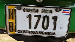Das Bild zeigt ein Autokennzeichen in Alajuela. Auf einem gelben, vertikalen Streifen links stehen die Buchstaben H und B untereinander. Auf der weißen Fläche steht in Schwarz Costa Rica, darunter 1701 und darunter Centroamerica. Rechts oben ist die costaricanische Flagge zu erkennen. 