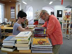 Das Bild zeigt eine Mitarbeiter der Mediathek, die an einem Tresen mit zahlreichen Büchern einen Mann bedient.