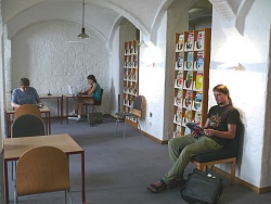 Das Bild zeigt den Zeitschriftenbereich der Mediathek Lahr unter einer weißen Gewölbedecke. Vor den Zeitschriftenregalen stehen kleine Tische und Stühle. Zwei Frauen und ein Mann sitzen dort und lesen Zeitschriften beziehungsweise arbeiten am Laptop.