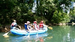 Das Bild zeigt sechs Menschen mit Schwimmwesten und Helmen in einem Schlauchboot auf einem Fluss bei Alajuela paddelnd. Am Ufer stehen Büsche und Bäume.