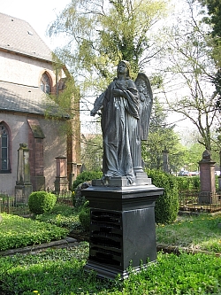 Das Bild zeigt eine Grabstatue auf dem Friedhof Stiftskirche in Lahr. Auf einem schwarzen Sockel steht an eine Säule gelehnt ein steinerner Engel mit nach oben gerichtetem Blick.