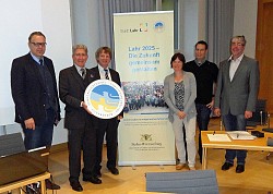 Das Bild zeigt die Übergabe des Siegels, das die Stadt Lahr für ihr Handlungskonzept Lahr 2025 erhalten hat. Zu sehen sind von links der erste Bürgermeister Guido Schöneboom und Oberbürgermeister Dr. Wolfgang Müller mit dem Siegel sowie vier weitere Personen um ein Banner mit der Überschrift "Lahr 2025 - Zukunft gemeinsam gestalten" gruppiert.