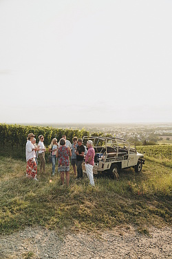 Gruppe von Menschen steht neben einem offenen Geländewagen auf einer Wiese mit Weinbergen im Hintergrund