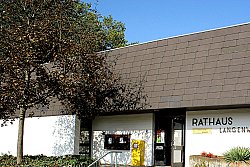 Das Bild zeigt das Rathaus in Langenwinkel, ein eingeschossiges weißes Gebäude mit nach hinten geneigtem schwarzen Pultdach. Links neben dem Eingang zu dem einige breite Stufen führen vor denen ein Blumenkübel steht befindet sich ein gelber Post-Briefkasten, rechts vom Eingang ein weiterer Blumenkübel. Rechts des Gebäudes stehen Büsche, links davor ein Baum.