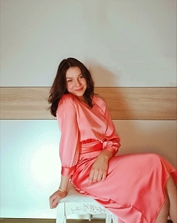 Die Künstlerin trägt ein lachsfarbenes, langes Kleid und sitzt schräg auf einem Stuhl. Sie blickt die Kamera glücklich an.