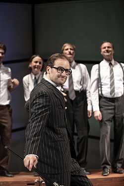 Ein Mann mit schwarzem Nadelstreifen-Anzug, dunklen zurück-gekämmten Haaren und einem schwarz glänzendem Gehstock lächelt süffisant in die Kamera. Im Hintergrund stehen mehrere lächelnde Personen mit schwarzen Hosen, Hosenträgern und weißen Hemden.