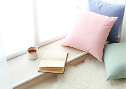 Ein Platz am Fenster, dekoriert mit drei Kissen einem weißen Schaffell, einem aufgeschlagenem Buch und einer Tasse Tee. 