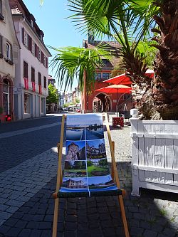 Das Foto zeigt einen Klappliegestuhl, der mit bunten Sehenswürdigkeiten aus Lahr bedruckt ist. Der Liegestuhl steht auf einer Straße. Im Hintergrund sind Blumen und ein großes Gebäude zu sehen. 