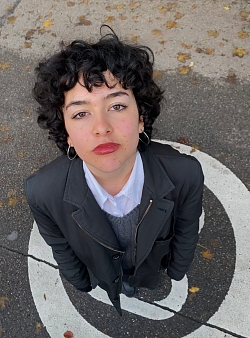 Die Kamera ist von oben auf die Künstlerin gerichtet, was das Bild in einer Vogelperspektive erscheinen lässt. Sie blickt die Kamera etwas streng direkt an.