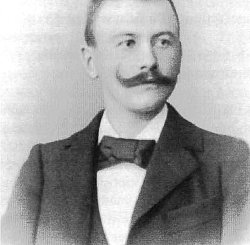Schwarz-weiß Fotografie von Ludwig Sütterlin. Er trägt einen seitlich nach oben gezwirbelten Schnauzbart, ein weißes Hemd, schwarzen Anzug und eine schwarze Fliege.