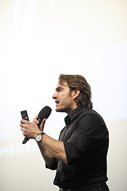 Der Referent ist von der Seite aufgenommen. Er spricht in ein schwarzes Mikro und hält einen Laserpointer in der anderen Hand. Er trägt ein schwarzes Hemd und hat halblanges durchgestuftes Haar. 