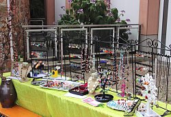 Das Bild zeigt einen Stand beim Kunsthandwerkermarkt. Er besteht aus einem Bierzelttisch mit einer hellgrünen Tischdecke, darauf ist eine Vielzahl an Schmuck an verschiedenen Ständern und auf Platten oder in Schachteln arrangiert.