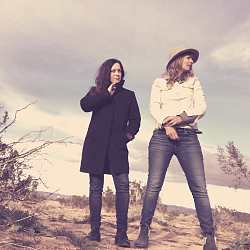 Zu sehen sind die zwei Sängerinnen Madion Violet. Sie stehen auf einer Düne, das Bild ist in einem Sepia-Farbton gehalten. Die Frau links hat dunkle Haare und trägt einen langen schwarzen Mantel. Die blonde Frau rechts trägt Jeans, ein weißes Hemd und einen Cowboy-Hut. Sie blickt nach rechts.