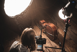 Ein Kameramann kniet vor einem Cello auf dem Boden mit einer Kamera. Das Cello ist von zwei Scheinwerfern beleuchtet. 