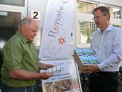 Zu sehen ist Manfred Kaiser, der Edwin Fischer Klimasparbücher übergibt. Beide sind sommerlich gekleidet und stehen vor dem Rathaus.