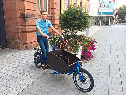 Das Bild zeigt Martin Stehr vom Lahrer Stadtplanungsamt vor dem Rathaus 2, ehemalige Luisenschule, auf einem bleuen Lastenrad sitzend. Er trägt ein blau-weiß kariertes Hemd und eine blaue Jeans. Im Hintergrund ein bunt bepflanzter Blumenkübel.
