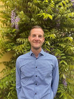 Portait eines Mannes mit kurzen blonden Haaren, lächelnd, gekleidet mit einem blauen Hemd.