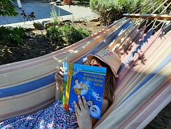 Aktion Heiss auf Lesen der Mediathek Lahr mit einem kind in der Hängematte, das ein Buch vor die Nase hält und Sonnenhut und Sonnenbrille trägt.