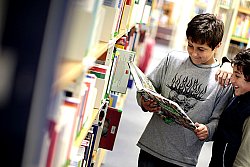 Zwei Jungs stehen zwischen Bücherregalen in der Mediathek. Einer hält ein aufgeschlagenes Buch in der Hand, in das beide lachend hineinsehen.