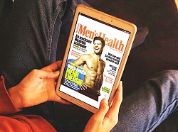 Das Bild zeigt eine sitzende Person. Man sieht nur einen kleinen Teil des Oberkörpers, die Hände und die Beine sowie einen Fuß. Die Person trägt einen roten Pullover, eine Jeans und keine Schuhe. In den Händen hält sie ein Tablet; auf dem Display ist die Online-Ausgabe der Zeitschrift 'Men's Health' zu sehen.
