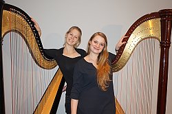 Die Harfenistinnen Mirjam Weschle und Stephanie Wunsch sind mit Ihren Harfen zu sehen. Beide Frauen haben blondes langes Haar und tragen eine schwarze Bluse und Hose. Sie haben jeweils einen Arm auf ihre Harfe gelegt und lächeln in die Kamera.