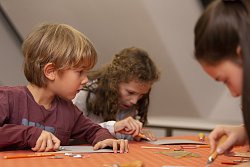 Drei Kinder, ein Junge und zwei Mädchen, sitzen an einem Tisch und malen oder basteln. Der Junge schaut gerade zu den anderen. Die beiden Mädchen sind konzentriert bei ihrer Arbeit.