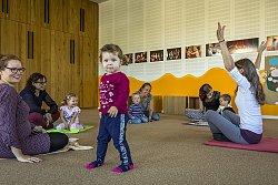 In einem Raum der Städtischen Musikschule sitzen Mütter mit ihren Kleinkindern auf dem Fußboden. Auch die Lehrerin sitzt dabei und hebt die Hände hoch in die Luft. Mütter und Kinder schauen sie lachend an. Ein Mädchen steht im Vordergrund und schaut verschmitzt in die Kamera. 