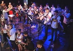 Zu sehen sind zahlreiche Musikerinnen und Musiker, die verschiedenen Blasinstrumente spielen. Vor ihnen sind Notenständer aufgestellt. Sie stehen auf einer Bühne und werden von einem Mann dirigiert.