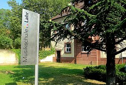 Ansicht des Gebäudes der Städtische Musikschule Lahr, im Vordergrund ein großes Schild mit der Aufschrift &#34;Städtische Musikschule Lahr&#34; und dem Logo der Stadt. Rechts im Bild ein großer Nadelbaum.