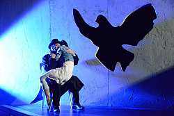 Frau mit weißem Kleid und Jeansjacke sitzt auf dem Schoß eines Mannes, der mit scharzem Mantel und Hut gekleidet ist, und drückt sich an ihn. In der hinteren Wand ist die Silhouette einer riesigen Taube herausgeschnitten. Szenerie ist mit blauem Licht bestrahlt.