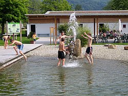 Zusehen sind drei Jungs an einer Wasserfontaine im Naturbad Sulz. Das Wasser kommt aus einem großen Stein. Die Jungs bespritzen sich gegenseitig. Im Hintergrund sitzen einige Badbesucher unter einer Überdachung vor dem Hauptgebäude. Rechts im Bild sind drei weitere Kinder zu sehen.