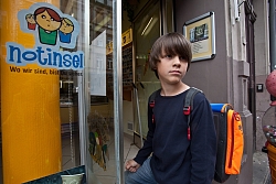 In der rechten Bildhälfte befindet sich ein Junge im Alter von ca. 9 Jahren. Er trägt einen Schulranzen auf dem Rücken und steht vor dem Eingang eines Geschäftes mit Schaufenster. Auf dem Schaufenster sieht man ganz groß den &#34;Notinsel&#34;-Sticker. 