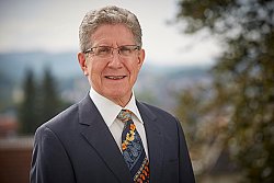 Das Bild zeigt Oberbürgermeister Dr. Wolfgang G. Müller.Er hat graue, kurze Haare, trägt eine eckige, im unteren Bereich rahmenlose Brille, einen grauen Anzug, ein weißes Hemd und eine Krawatte mit Mustern und gelben und orangen Blüten.