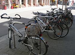 Das Bild zeigt silberne Fahrradanlehnbügel an denen mehrere Fahrräder lehnen an der Kaiserstraße auf der gegenüberliegenden Straßenseite ist das Haus Stoesser-Fischer zu erkennen