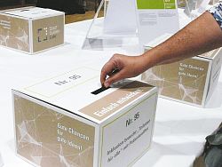 Zu sehen ist eine weiße Abstimmungsbox und eine Frauenhand, die einen Stadtgulden hinein wirft.